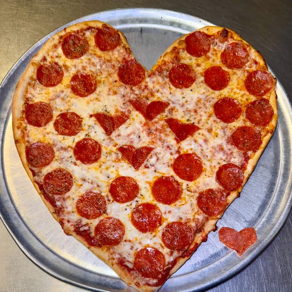 Heart-shaped pepperoni pizza with melted cheese and a small pepperoni heart on a metal tray.