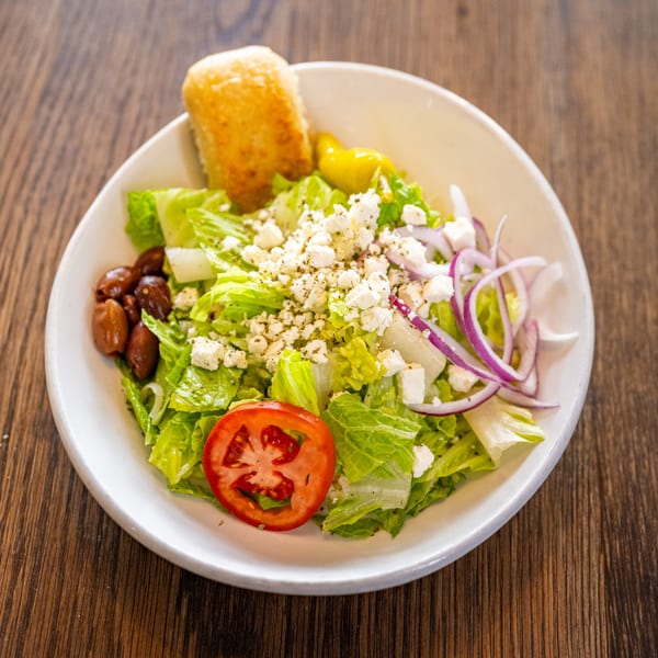 A fresh Greek salad with lettuce, feta, olives, red onion, tomato, and a breadstick.