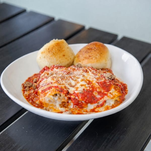 A white bowl of lasagna with melted cheese, red sauce, ground meat, and two bread rolls.