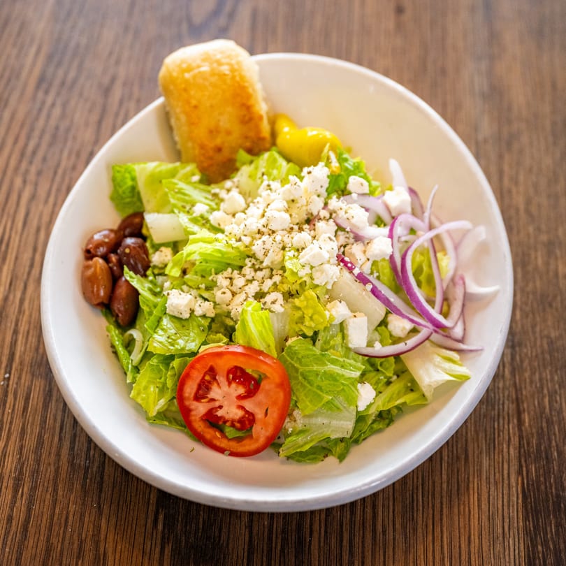 A fresh Greek salad with lettuce, feta, olives, red onion, tomato, and a breadstick.