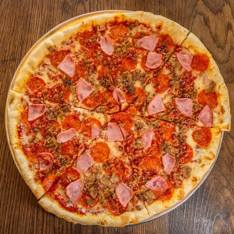 A whole meat lover's pizza, topped with pepperoni, ham, and ground meat, on a wooden table.