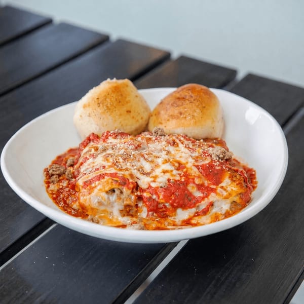 A white bowl of lasagna with melted cheese, meat sauce, and two bread rolls on a dark wooden table.