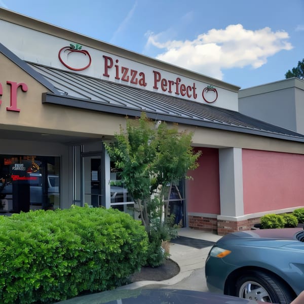 The exterior of Pizza Perfect restaurant, with its red sign and tomato logo, on a sunny day.