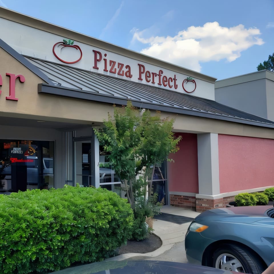 The exterior of Pizza Perfect restaurant, with its red sign and tomato logo, on a sunny day.