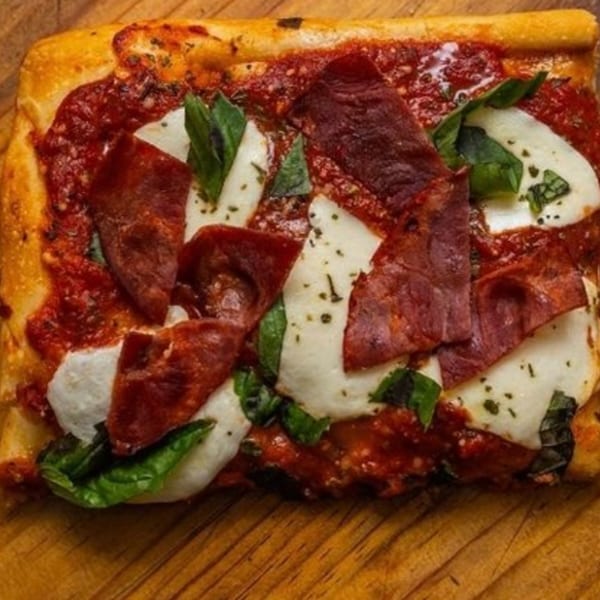 A rectangular slice of pizza with tomato sauce, mozzarella, pepperoni, and fresh basil on a wooden board.