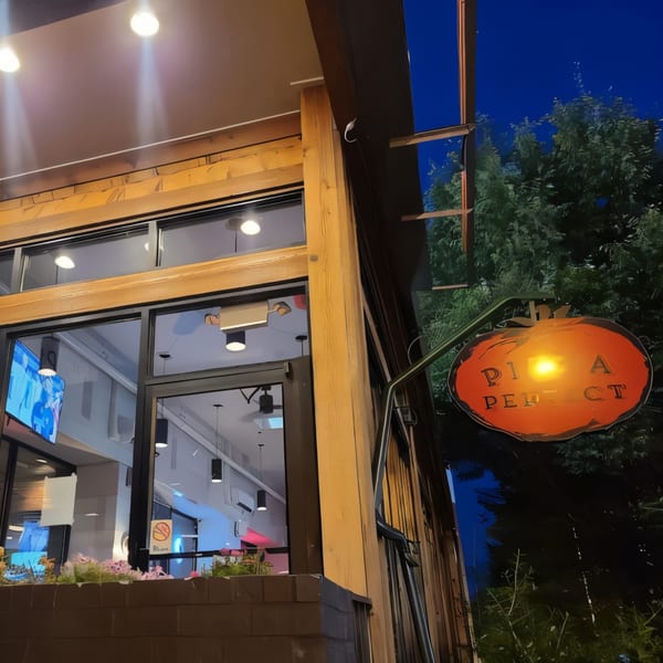 Exterior of "Pizza Perfect" restaurant at dusk, with a glowing orange sign and visible interior.